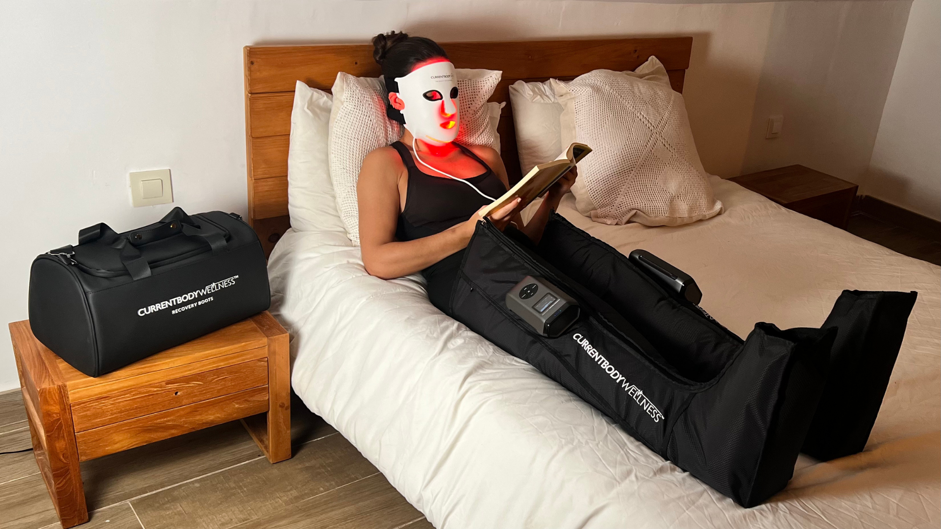 Carmen Bauzá usando una máscara LED facial de luz roja y botas de presoterapia CurrentBody en casa, leyendo en la cama.